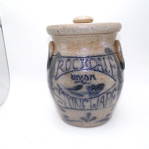 May include: A brown and blue stoneware crock with a lid. The crock has a blue inscription that reads "Rockdale Union Stoneware".