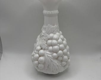 Puede incluir: Un jarrón blanco de estilo vintage, con una parte superior con volantes y un cuerpo bulboso. El jarrón está decorado con un patrón en relieve de uvas y hojas, añadiendo un toque de elegancia clásica. El jarrón está hecho de un material brillante y opaco.
