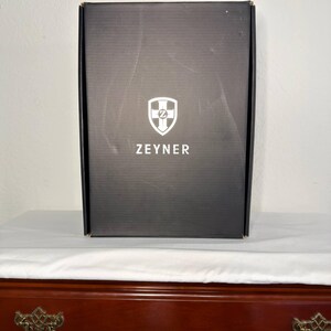 May include: A black rectangular box with the word "ZEYNER" printed in white below a white and blue shield logo. The box is sitting on a white surface, and the background is a light wall.
