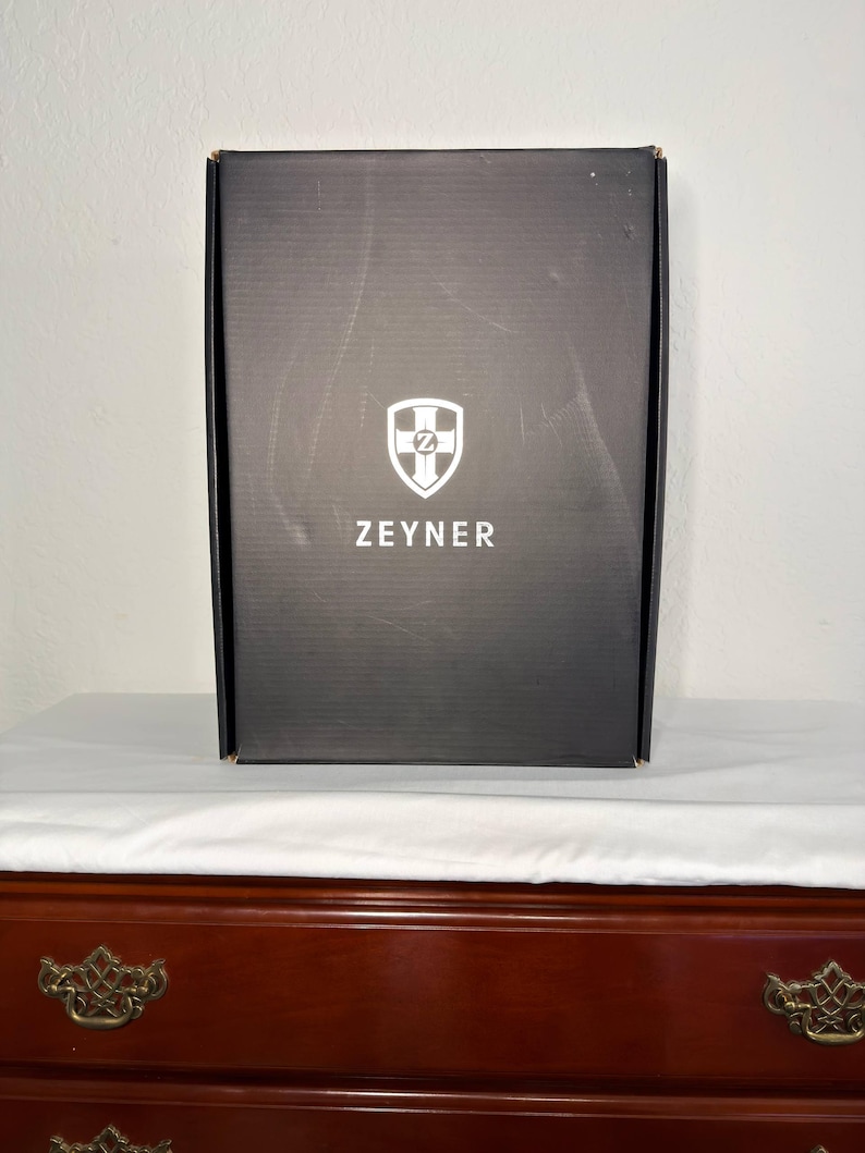 May include: A black rectangular box with the word "ZEYNER" printed in white below a white and blue shield logo. The box is sitting on a white surface, and the background is a light wall.