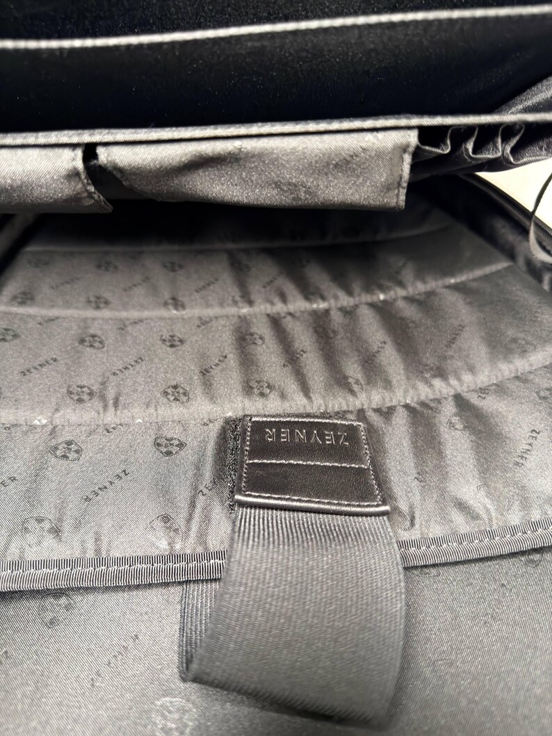 May include: Black interior of a bag with a fabric lining. The lining has a repeating pattern. A black strap with the word "ZEYNER" is sewn into the bag.