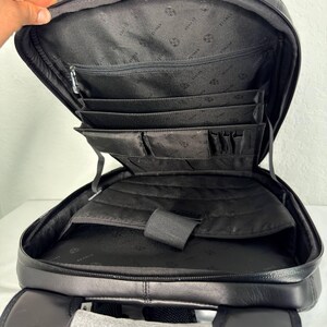 May include: Black leather backpack with multiple interior compartments, including a zippered pocket and several smaller pockets for organization. The backpack has a padded back and adjustable straps. The interior lining features a repeating pattern.