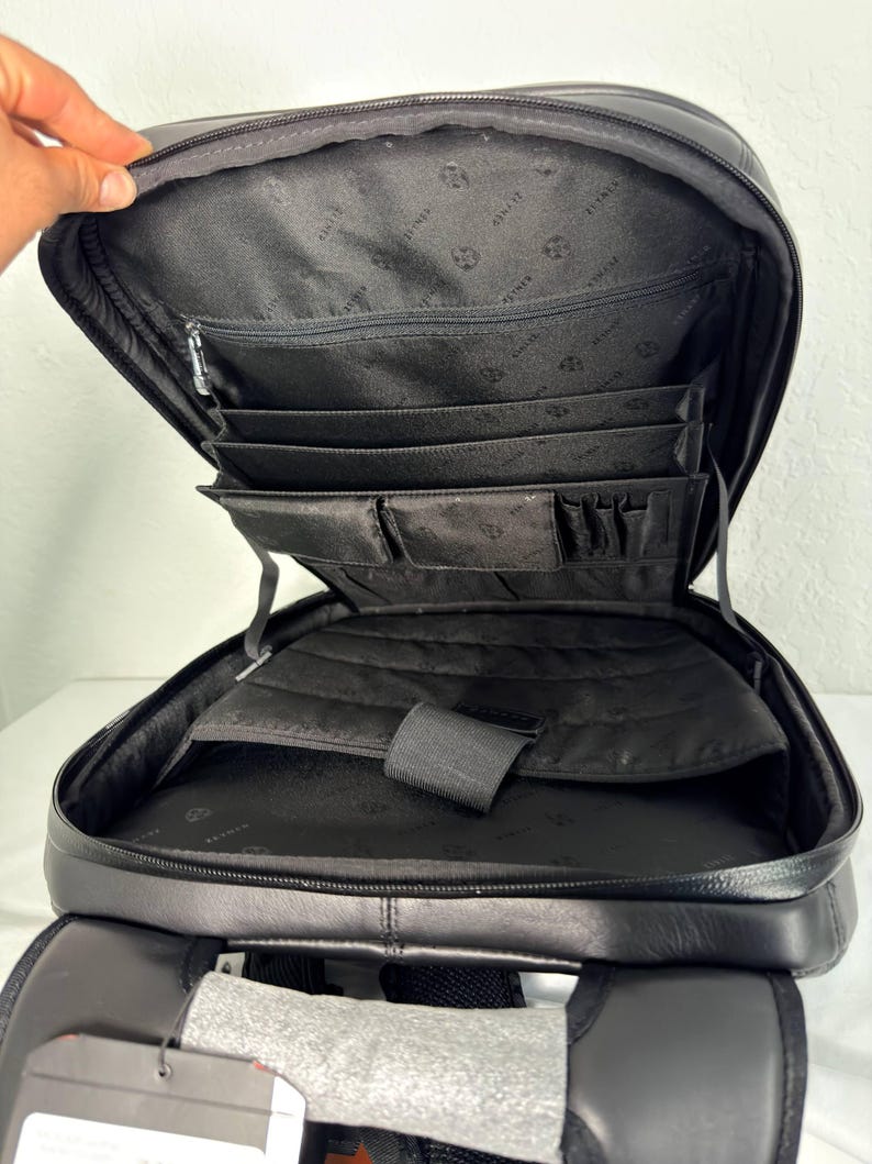 May include: Black leather backpack with multiple interior compartments, including a zippered pocket and several smaller pockets for organization. The backpack has a padded back and adjustable straps. The interior lining features a repeating pattern.
