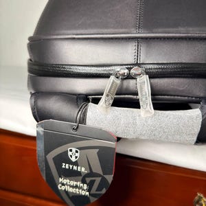 May include: Black leather travel bag with a zipper closure and a tag that reads "Zeyner Motoring Collection". The bag has a structured design and appears to be made of high-quality materials. The bag is designed for travel.