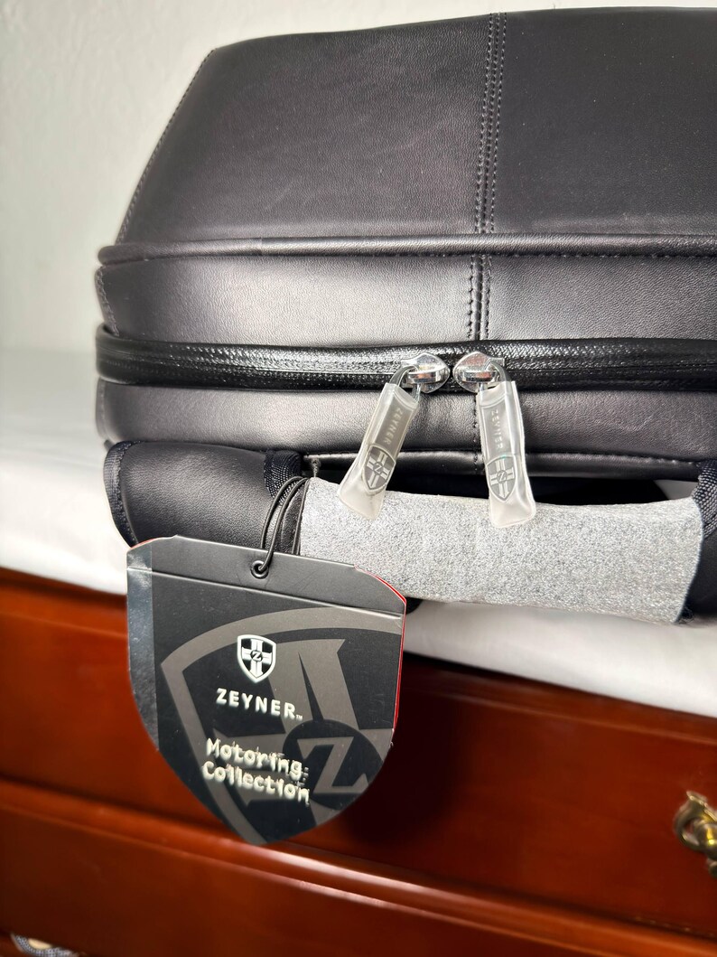 May include: Black leather travel bag with a zipper closure and a tag that reads "Zeyner Motoring Collection". The bag has a structured design and appears to be made of high-quality materials. The bag is designed for travel.