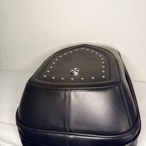 May include: Black leather backpack with a rounded top and silver-colored rivets. The backpack features a silver emblem and two zippers at the bottom. The bag has a structured design and appears to be made of high-quality materials.