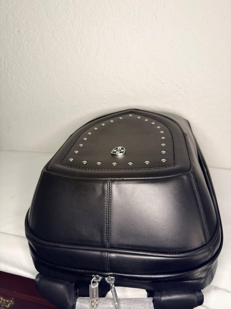 May include: Black leather backpack with a rounded top and silver-colored rivets. The backpack features a silver emblem and two zippers at the bottom. The bag has a structured design and appears to be made of high-quality materials.