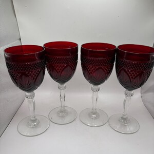 May include: Four ruby red wine glasses with clear stems and bases. The glasses have a cut glass design on the bowl. The stems are clear and the bases are round. The glasses are arranged in a row.