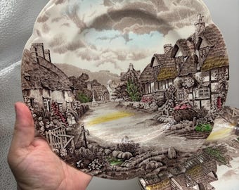 Pode incluir: Um conjunto de pratos decorativos com uma paisagem de vila. Os pratos apresentam casas, árvores e um céu nublado, com uma borda ondulada. As cores principais são castanho, bege e azul.