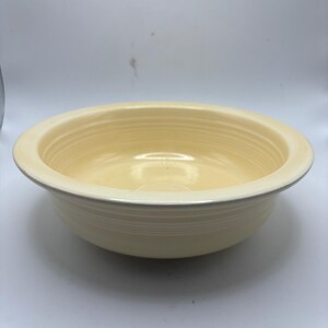 May include: A pale yellow ceramic bowl with a wide rim and a slightly textured surface. The bowl has a simple, classic design, suitable for serving or display. The bowl is a light cream color.