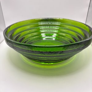 Könnte beinhalten: Eine grüne Glasschale mit einem strukturierten, gerippten Design. Die Schale ist kreisförmig und hat eine breite, flache Form. Die Farbe ist ein leuchtendes, transparentes Grün, und die Schale scheint aus dickem Glas zu bestehen.