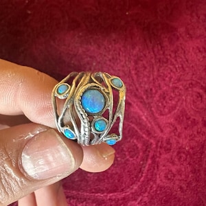 Puede incluir: Un anillo de plata con una piedra central de ópalo azul ovalada, rodeada de pequeños detalles de ópalo azul. El anillo tiene un diseño abierto y artístico con una banda texturizada. El anillo se sostiene sobre un fondo rojo.