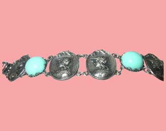 Op de afbeelding: Een zilveren armband met ovale bedels met een vrouwenprofiel en turquoise glazen stenen.