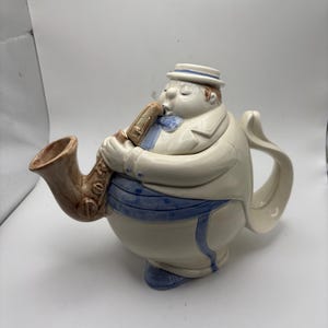 May include: A whimsical teapot shaped like a musician playing a saxophone. The ceramic teapot is off-white with blue and brown accents. The musician wears a hat and a bow tie. The saxophone's bell serves as the teapot's spout.