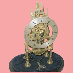 Può includere: Un orologio scheletrico intricato color oro con numeri romani sul quadrante. L'orologio è su una base nera con una chiave di carica.