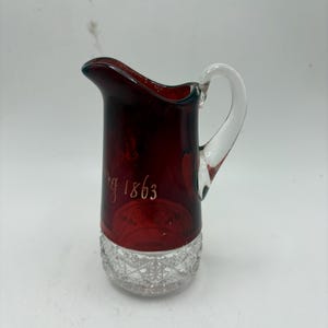 May include: A ruby red glass jug with a clear handle and a diamond-cut base. The jug has the etched text "g 1863" on the front. The jug is likely vintage and suitable for serving beverages.