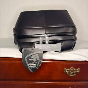 May include: Black leather bag with a unique, geometric design. Features a silver zipper and a tag that reads "Zeyner Motoring Collection". The bag is placed on a white surface, with a wooden dresser in the background.