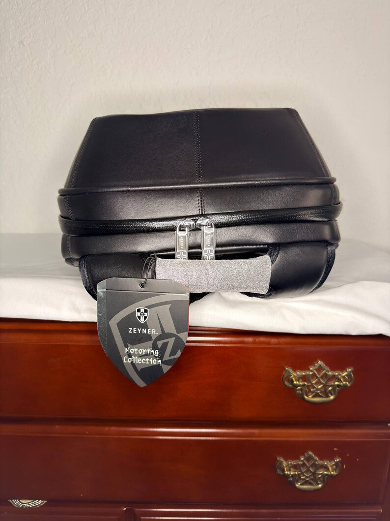 May include: Black leather bag with a unique, geometric design. Features a silver zipper and a tag that reads "Zeyner Motoring Collection". The bag is placed on a white surface, with a wooden dresser in the background.