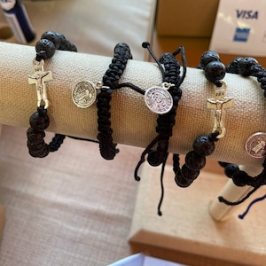 May include: Black lava stone bracelets with silver-tone religious charms, including crucifixes and medallions. The bracelets are displayed on a beige fabric-covered stand, showcasing their intricate designs and spiritual theme.