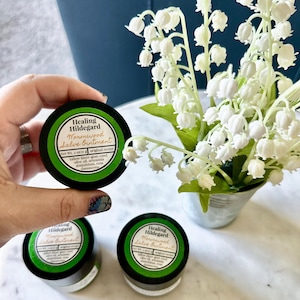 Könnte beinhalten: Drei kleine grüne und schwarze Gläser mit Wormwood Salve Ointment mit dem Text "Healing Hildegard" und den aufgelisteten Zutaten auf dem Etikett. Die Gläser stehen auf einer weißen Oberfläche mit einem Strauß weißer Maiglöckchen in einem Metalltopf.