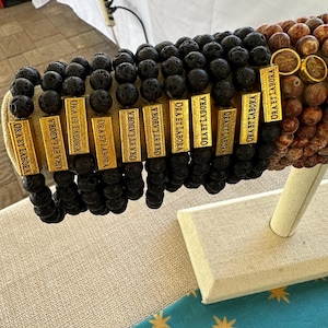 May include: A display of black lava rock bracelets with gold charms that say "Ora Et Labora".