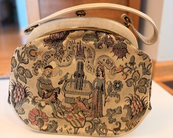 May include: A structured handbag featuring a cream-colored frame and handle with a gold-tone metal clasp. The body is made of woven tapestry fabric depicting a medieval scene with a central fountain, two figures, and floral motifs in muted tones.