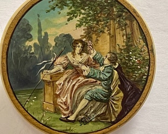 May include: A round, ivory-colored box with a painted scene of a couple in a garden setting. The woman is wearing a pink dress and the man is wearing a blue jacket. The scene is surrounded by a gold border.