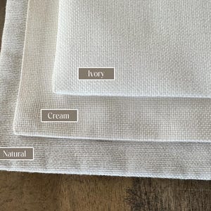 May include: Three fabric swatches in different shades of white: natural, cream, and ivory. The swatches are stacked on top of each other, with the natural swatch at the bottom and the ivory swatch at the top.