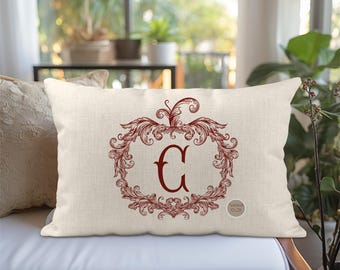 May include: A rectangular throw pillow in a natural beige fabric featuring a dark red ornate scrollwork frame centred around the letter G. A small circular tag on the lower right corner displays the text Natural 12x20.