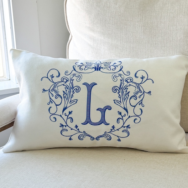 Puede incluir: Cojín rectangular color crema con un diseño bordado en azul. El diseño presenta la letra "L" en una fuente decorativa, rodeada por una corona floral ornamentada. El cojín está colocado sobre una silla de tono neutro.