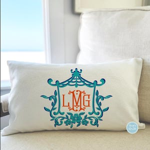 May include: A white pillow with a teal and orange monogram design. The monogram is in a decorative frame with a teal pagoda-like structure at the top. The pillow is 12x20 inches and is labeled "Ivory".