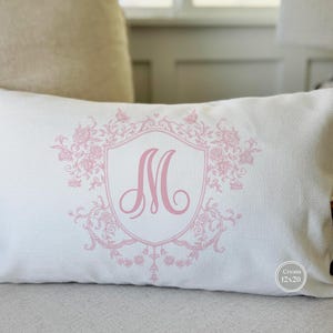 May include: Cream-colored rectangular pillow with a pink floral crest design and the letter "M". The pillow measures 12 x 20 inches. The design includes a shield shape with floral accents.