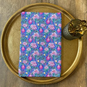 May include: A blue and pink floral patterned cloth napkin rests on a gold-colored tray. The napkin features a repeating pattern of pink ornaments, white flowers, and green leaves. A small container of gold forks sits on the tray.
