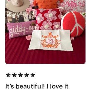 May include: A white pillow with a pink and orange monogram of the letters "JCH" in a decorative frame. The pillow is on a bed with a pink and white striped bedspread and several other pillows, including a pink pillow with white flowers, a red pillow with a peace sign, and a pink pillow with the words "giddy-up" embroidered on it.