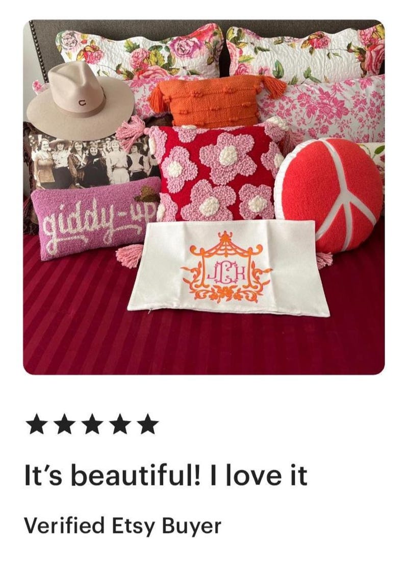 May include: A white pillow with a pink and orange monogram of the letters "JCH" in a decorative frame. The pillow is on a bed with a pink and white striped bedspread and several other pillows, including a pink pillow with white flowers, a red pillow with a peace sign, and a pink pillow with the words "giddy-up" embroidered on it.