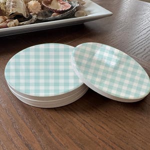 May include: A stack of round coasters and one individual coaster featuring a light blue and white gingham check pattern. The coasters have a smooth, glossy top surface and a thick, matte white base, displaying a consistent checkered design.