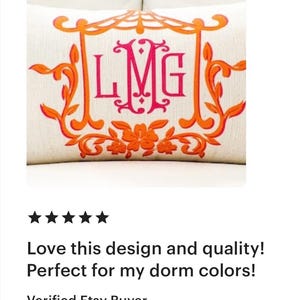 May include: A white pillow with an orange and pink monogram of the letters "LMG" in a decorative frame. The pillow is embroidered with floral details.