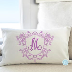 May include: Ivory rectangular pillow with a lavender floral crest design and the letter "M". The pillow is 12x20 inches and rests on a neutral-toned surface. The design includes floral elements and a decorative border.