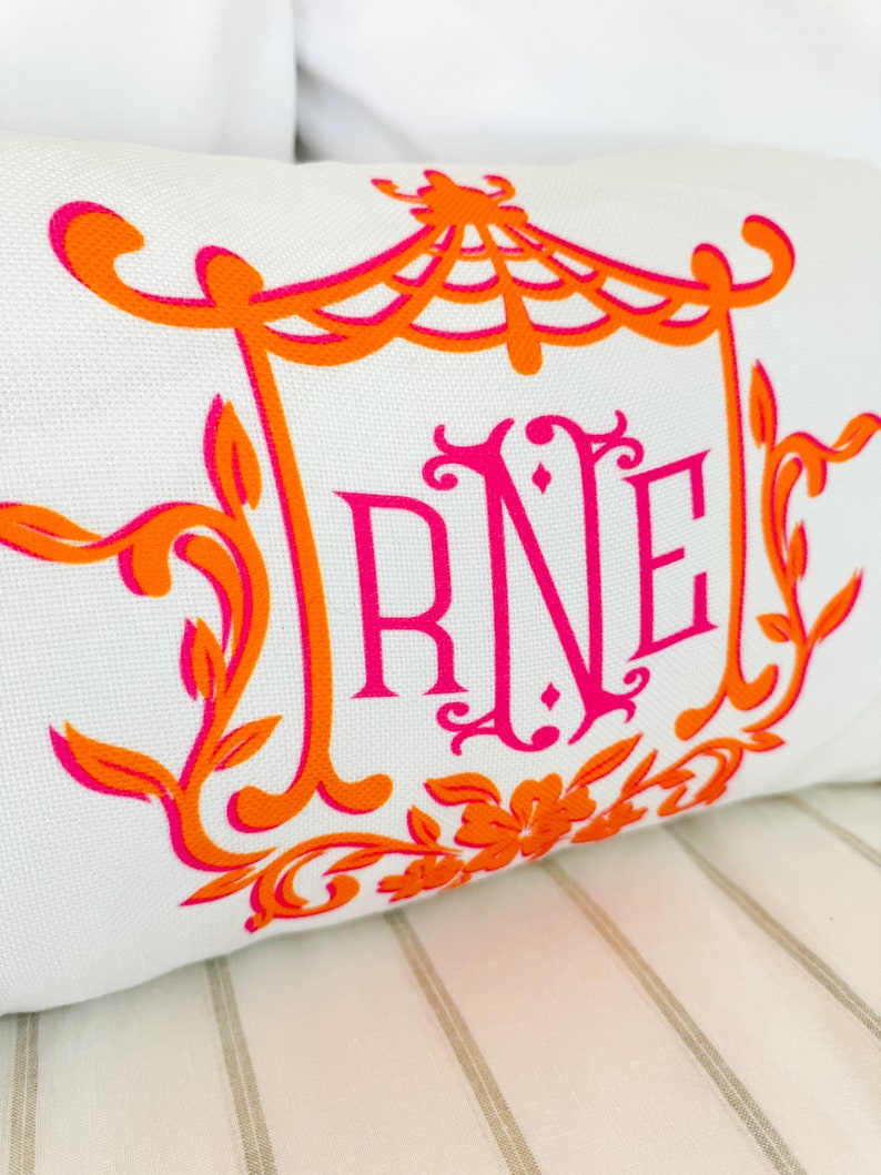 May include: A white pillow with an orange and pink monogram design. The monogram is "RNE" and is surrounded by a decorative frame with a floral design.
