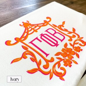 May include: A white canvas tote bag with an orange and pink monogram design. The monogram is in a decorative frame with floral accents. The text "TOB" is embroidered in pink.
