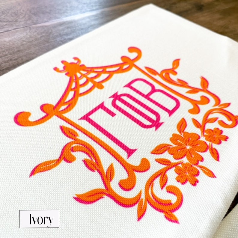 May include: A white canvas tote bag with an orange and pink monogram design. The monogram is in a decorative frame with floral accents. The text "TOB" is embroidered in pink.