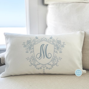May include: A white pillow with a blue embroidered monogram "M" in a decorative frame with floral accents. The pillow is 12x20 inches.