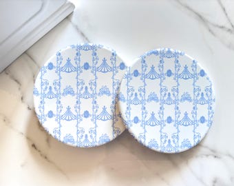 May include: Two round white coasters featuring a repeating light blue pattern of vertical lines, floral motifs, and arched shapes. The coasters have a smooth, flat surface and are arranged side by side, displaying a consistent design across both items.