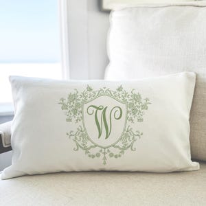 May include: A rectangular white throw pillow featuring a centered sage green graphic design. The design consists of a shield-shaped crest containing the letter W, surrounded by intricate floral and scrollwork patterns in a matching green color.