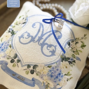 May include: A white ring pillow with blue floral designs and a blue ribbon. The pillow has a blue crest with the letter "M" in the center and the text "# 2023" at the bottom.