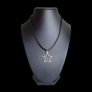 May include: A silver star pendant necklace on a black cord. The star is a simple outline design. The necklace is displayed on a black mannequin against a black background.