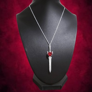 Pode incluir: Um colar de prata com um pingente de espada com uma pedra preciosa vermelha em forma de coração.