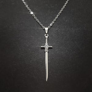 May include: A silver sword pendant necklace with a detailed design on the hilt and blade. The necklace is on a silver chain.