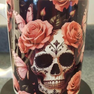 May include: White ceramic mug featuring a sugar skull design with pink roses and butterflies. The skull has intricate black detailing and is surrounded by floral and insect elements. The mug has a white handle and rim.