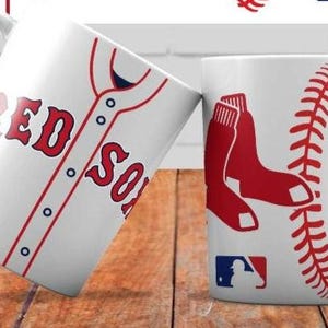 May include: Two white ceramic coffee mugs with baseball-themed designs. One mug features a red and blue baseball jersey with the text "RED SOX". The other mug features a red baseball glove and a red baseball stitch design.